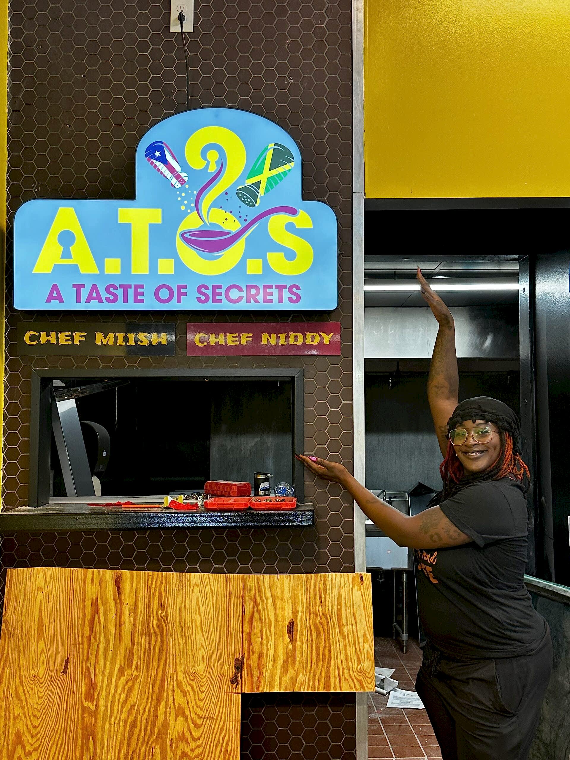 Illuminated light box sign for A Taste of Secrets in Atlanta, GA, featuring bright, colorful branding above the service window in a busy market setting